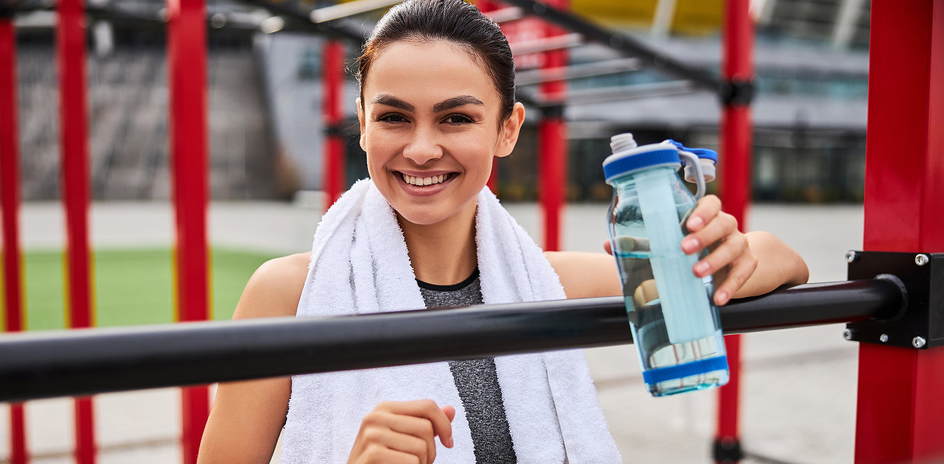 Woman with a towel around her neck holding a water bottle on outdoor gym equipment