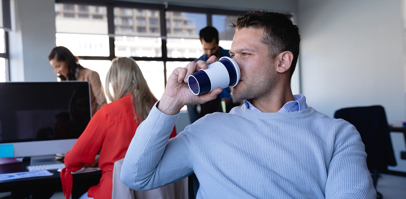 Man drinking from a coffee cup in an office setting with colleagues in the background.